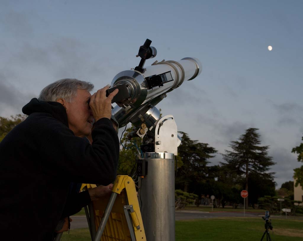 An amateur astronomer at the eyepiece of a telescope on an equatorial mount at dusk, with the Moon visible in the sky.